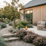 A serene Scandinavian-style garden photographed in soft natural light, featuring a simple light wood deck terrace with clean lines, surrounded by ornamental grasses gently swaying in the breeze, clusters of heather in muted purple tones, and a slender rowan tree with bright red berries. Natural stone steps and a gravel pathway wind through the space, bordered by low-maintenance perennials arranged in calm, repeating groups. Warm Edison-style outdoor lanterns cast a gentle amber glow near comfortable wooden furniture with soft textiles. The garden seamlessly connects to a Scandinavian-style home facade, with the same natural materials echoed throughout. The overall palette is earthy and understated — pale wood, grey stone, silver-blue grass blades, and muted greens — conveying a sense of peaceful minimalism, hygge coziness, and harmony with nature. Editorial photography style, shallow depth of field, golden hour lighting.